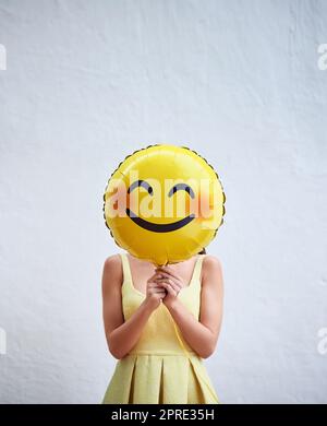 Lächeln Sie immer wieder. Studioaufnahme einer unkenntlichen Frau, die vor grauem Hintergrund einen lächelnden Emoticon-Ballon vor ihrem Gesicht hält. Stockfoto