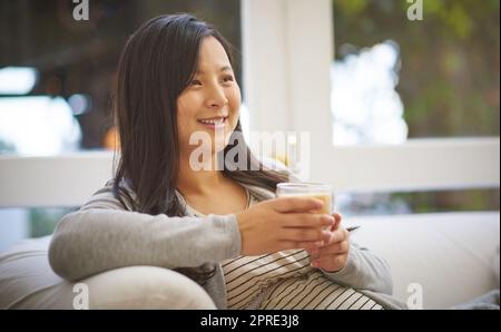 Ich kann es kaum erwarten, meinen kleinen Jungen zu treffen. Eine attraktive junge Schwangere, die einen Eiskaffee trinkt, während sie sich zu Hause auf dem Sofa entspannt. Stockfoto