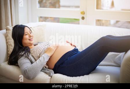 Ich kann es kaum erwarten, Sie in meinen Armen zu halten. Eine attraktive junge Schwangere, die sich zu Hause auf dem Sofa entspannt. Stockfoto