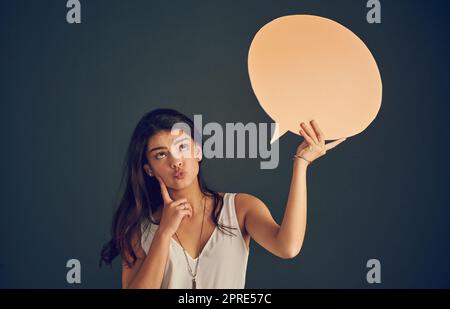 Lassen Sie mich kurz nachdenken. Studioaufnahme einer sorglosen jungen Frau, die eine Sprechblase hält, während sie sich Gedanken macht und vor einem dunklen Hintergrund steht. Stockfoto