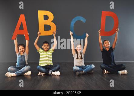 Jetzt kenne ich mein ABC. Studioportrait einer vielfältigen Gruppe von Kindern, die Buchstaben des Alphabets vor einem grauen Hintergrund hochhalten. Stockfoto