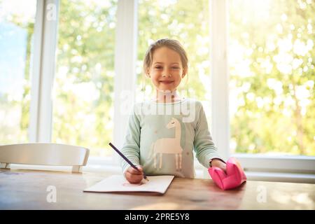 Zeichnen ist eines meiner liebsten Dinge zu tun. Porträt eines fokussierten kleinen Mädchens, das Bilder in einem Buch zeichnet, während es zu Hause auf die Kamera schaut. Stockfoto