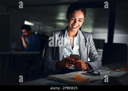 Frau, die spät am Telefon SMS liest, eine Nachricht lächelt und sich online Beiträge in sozialen Medien ansieht, während sie nachts in einem dunklen Büro arbeitet. Zufriedener Mitarbeiter, der E-Mails liest und Überstunden macht Stockfoto