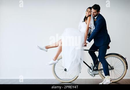 Auf in unsere Flitterwochen. Studioaufnahme eines frisch verheirateten jungen Paares, das vor grauem Hintergrund zusammen mit dem Fahrrad unterwegs ist. Stockfoto