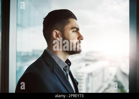 Sei ein Chef, baue ein Imperium. Ein junger Geschäftsmann, der nachdenklich aus einem Bürofenster schaut. Stockfoto