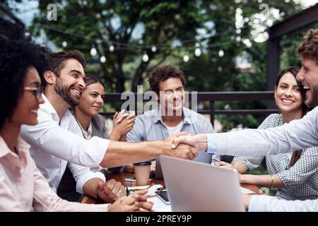 Heres an neue Geschäftspartner. Zwei hübsche junge Geschäftsleute, die sich die Hände schütteln, während sie mit ihren Kollegen im Freien sitzen. Stockfoto