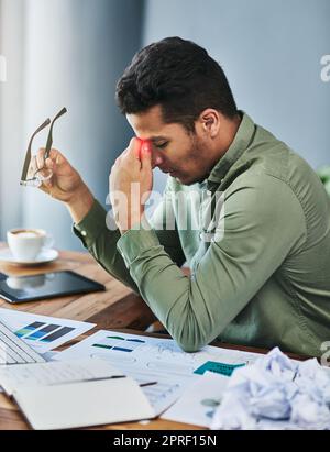 Diese Kopfschmerzen sind sehr irritierend. Ein unangenehmer junger Geschäftsmann, der seine Stirn mit schmerzhafter Hand hält, während er an seinem Schreibtisch im Büro sitzt. Stockfoto