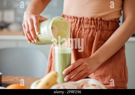 Frau, die einen gesunden Smoothie in ein Glas aus einem Mixbecher auf einer Theke zur Entgiftung eingießt. Eine Frau, die in ihrer Küche frischen grünen Fruchtsaft mit Gemüse und Verbrauchsmaterialien für einen gesunden Lebensstil herstellt. Stockfoto