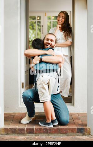 Liebevoller Vater umarmt und umarmt Sohn, Liebe vom Vater zum Sohn oder Eltern verabschieden sich von dem Kind auf der Veranda zu Hause. Eine glückliche Familie grüßt den kleinen Jungen mit der Mutter, die in der Tür oder im Hauseingang steht. Stockfoto