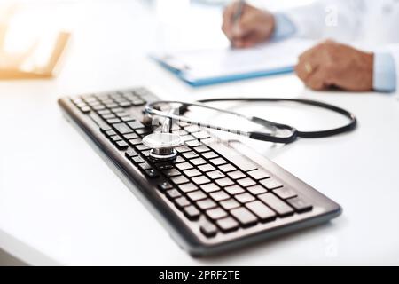 Neue Schule trifft auf alte Schule. Eine Tastatur mit Stethoskop darauf, während ein unkennbarer Mann auf einer Form im Hintergrund schreibt. Stockfoto