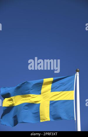 Die Nationalflagge Schwedens in den Farben Blau und Gelb im Wind auf einer Fahnenstange. Die Krone ist die Währung Schwedens. Der blaue Himmel dient als Hintergrund. Stockfoto