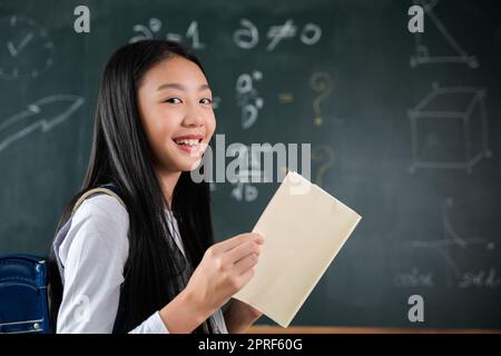 Glückliches, wunderschönes, asiatisches Schulmädchen steht mit Büchern vor der Tafel des Klassenzimmers Stockfoto