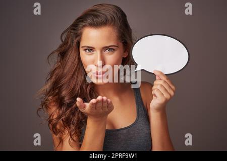 Liebe von den Lippen senden. Studioaufnahme einer attraktiven jungen Frau, die eine leere Sprechblase hält und einen Kuss auf einen grauen Hintergrund bläst. Stockfoto