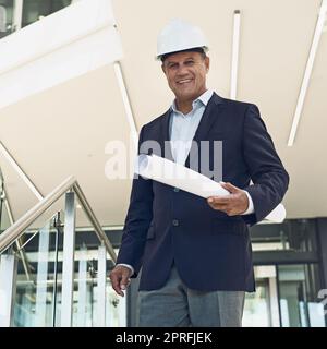 Ohne ihn wird es keine Entwicklung geben. Porträt eines fröhlichen, professionellen Architekten, der in die Kamera schaut, während er Baupläne in einem Gebäude hält. Stockfoto