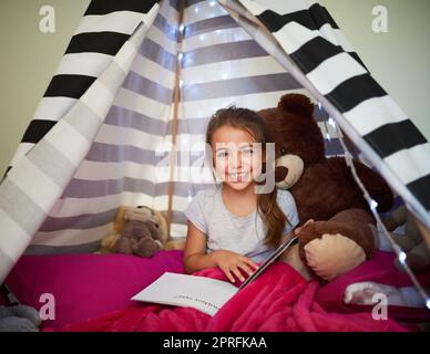 Es ist ihre eigene kleine Leseecke. Portrait eines kleinen Mädchens, das mit ihrem Teddybären zu Hause in einem Zelt ein Buch liest. Stockfoto