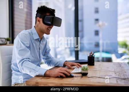 Eingetaucht in eine virtuelle Welt. Ein glücklicher junger Geschäftsmann, der ein Virtual-Reality-Headset trägt, während er an seinem Schreibtisch im Büro arbeitet. Stockfoto