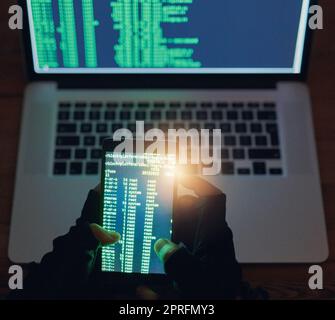 Durchbrechen mehrerer Netzwerke. Ein nicht erkennbarer Hacker, der im Dunkeln ein Handy und einen Laptop benutzt. Stockfoto