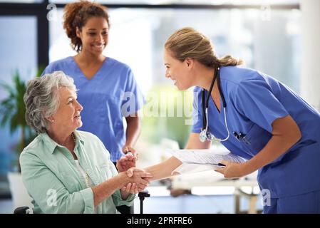 Ein Arzt, der Krankenakten hält und sich mit einer lächelnden älteren Frau, die in einem Rollstuhl sitzt, die Hände schüttelt. Stockfoto