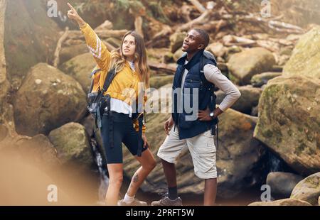 Fitness-Wandern, Natur und Paare beim Training, die gemeinsam nach Wegen suchen, um einen felsigen Pfad in der Natur zu nehmen. Mann und Frau aus verschiedenen Rassen zeigen auf Motivation und Navigation während der Wanderung. Stockfoto