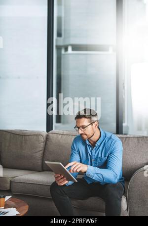 Ein Geschäftsmann, der sein digitales Tablet benutzt, während er auf einem Sofa im Büro sitzt. Stockfoto