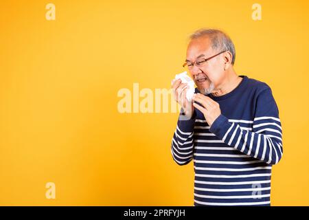 Porträt älterer alter Mann, der auf gelbem Hintergrund die Nase bläst, mit Tissuestudio-Schuss isoliert, älterer Mann aus Asien erkältet sich an Grippe und Niesen vor Krankheit Stockfoto