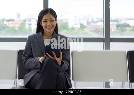 Junge asiatische Geschäftsfrau in grauem Anzug sitzt auf einer Bank in der Nähe des Fensters, verwenden Tablet-Computer auf der Suche nach interessanten Anlageinformationen. Stockfoto