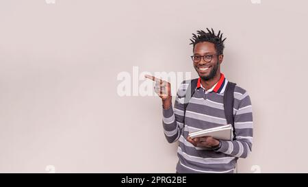 Porträt eines jungen afroamerikanischen Mannes mit kurzen Dreadlocks, Rucksack und Notizbüchern, lächelnd in die Kamera und zeigend auf Kopierraum. Das Studio wurde aufgenommen Stockfoto