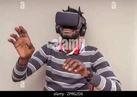 Afroamerikanischer Mann mit kurzen Dreadlocks, der ein Virtual-Reality-Headset mit integrierten Kopfhörern trägt, sich bewegende Hände und Gesten im Metaverse. Stockfoto