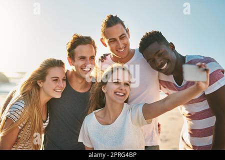 Die Saison für Selfies. Eine glückliche Gruppe von Freunden, die zusammen Selfies am Strand machen. Stockfoto