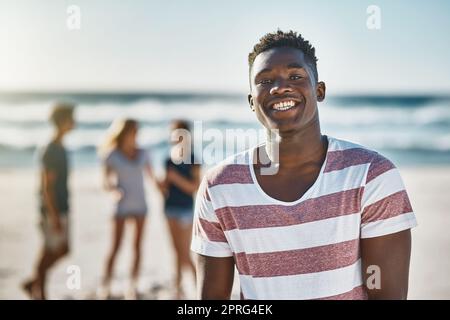 Wir kommen hierher, um unsere Dosis an Vitamin Meer zu erhalten. Porträt eines glücklichen jungen Mannes, der am Strand posiert, mit seinen Freunden im Hintergrund. Stockfoto