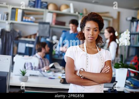 Shes eine ernsthafte Näherin. Porträt einer jungen Modedesignerin, die mit gefalteten Armen steht, während ihre Kollegen im Hintergrund arbeiten. Stockfoto