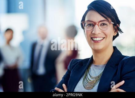 Manche nennen mich Visionärin. Eine Geschäftsfrau, die mit gekreuzten Armen und ihren Kollegen im Hintergrund steht. Stockfoto