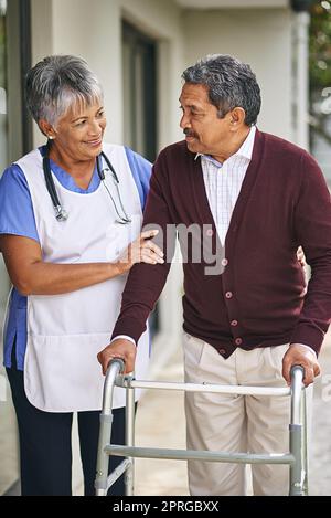 Eine Krankenschwester, die mit ihrem älteren Patienten und seinem Spaziergänger draußen spazieren geht. Stockfoto