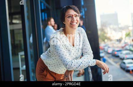 Die Zukunft sieht hell aus, von wo aus ich stehe. Eine junge Geschäftsfrau steht auf einem Balkon mit ihren Mitarbeitern im Hintergrund. Stockfoto