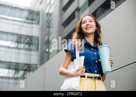 Asiatische schöne Geschäftsfrau zuversichtlich lächelnd mit Stofftasche hält Stahl Thermoskannen Becher Wasser Glas sie zu Fuß im Freien auf der Straße in der Nähe Modus Stockfoto