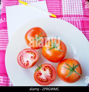 Frische reife rote Tomaten Tomaten in Scheiben geschnitten und mit Wassertropfen und einem grünen Stiel auf eine weiße Platte, neben einem Blatt Notepad schreiben und Steuerung Stockfoto