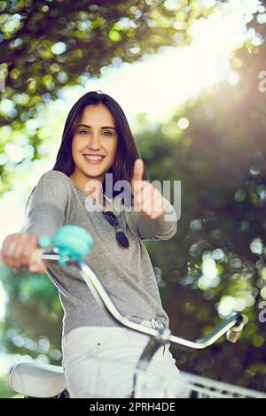 Treten Sie im Zweifelsfall in die Pedale. Porträt einer attraktiven jungen Frau, die den Daumen nach oben zeigt, während sie draußen mit dem Fahrrad fährt. Stockfoto