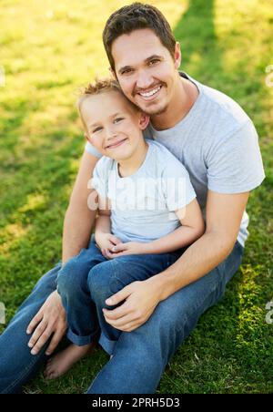 Gute Zeit mit Papa. Porträt eines lächelnden Vaters und seines kleinen Sohnes, der auf dem Gras in einem Park sitzt. Stockfoto
