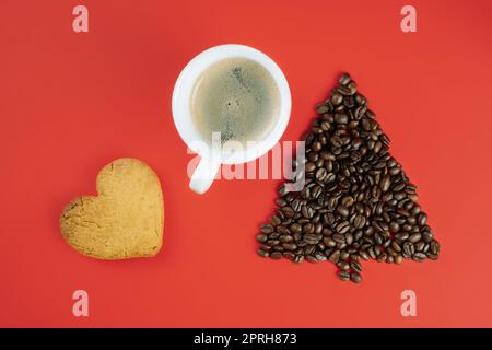 Braune Kaffeebohnen in Form eines Weihnachtsbaums mit einer Tasse Kaffee und einem herzförmigen Keks auf rotem Hintergrund. Flach liegend Stockfoto