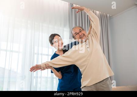 Der alte ältere Mann trainiert gerne bei einem Physiotherapeuten für ausgestreckte Arme zu Hause, der asiatische Physiotherapeut-Patient stillt ältere Menschen beim Trainieren Stockfoto