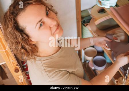 Lächelnde, fröhliche lockige Frau, Inhaberin des Geschäfts, die bunte Silikon-Babygeschirr aus hölzernen Regalen für Lieferauftrag wählt. Werkstatt, Arbeit zu Hause, Stockfoto