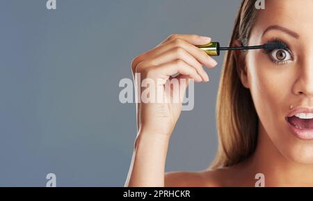 Covergirl Wimpern in der Herstellung. Studioportrait einer schönen jungen Frau, die vor grauem Hintergrund Mascara aufsetzt. Stockfoto