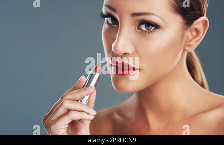 Mit dem roten Lippenstift kannst du deine Make-up-Routine aufpeppst. Studioportrait einer schönen jungen Frau, die auf einem grauen Hintergrund einen roten Lippenstift aufsetzt. Stockfoto