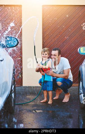 Ich liebe es, dass mein Vater mich in die Arbeit mit einbezieht. Die ganze Aufnahme eines Vaters und eines Sohnes, die mit einer Schlauchleitung spielen, während sie ein Auto gemeinsam waschen. Stockfoto