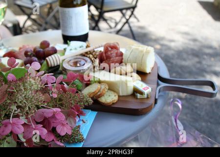 Weidebrett mit verschiedenen Käsesorten, Aufschnitt, Crackern, wne und Blumenstrauß zum Essen im Freien Stockfoto