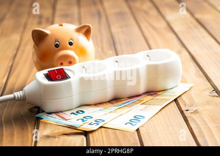 Steckdose, Sparschwein und Euro-Geld auf Holztisch. Konzept steigender Strompreise. Stockfoto