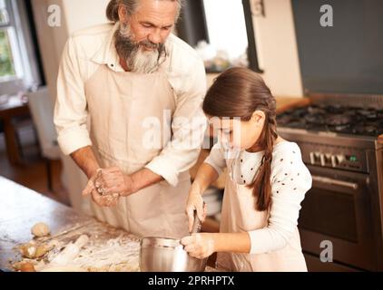 Opa lehrt sie ein oder zwei Dinge. Ein Mädchen, das sich mit ihrem Großvater verklebt, während sie in der Küche backen Stockfoto