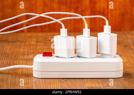 Die elektrische Verlängerungsleiste mit angeschlossenen weißen Netzsteckern auf dem Holztisch. Ladekonzept für verschiedene Geräte. Stockfoto