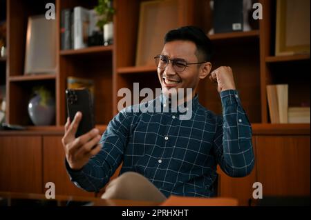 Glückliche asiatische Geschäftsfrau, die auf seinen Smartphone-Bildschirm blickt und feiert, nachdem sie eine gute E-Mail oder sms erhalten hat. Stockfoto