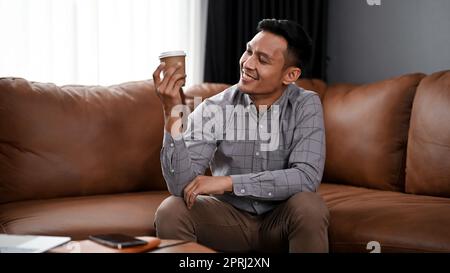Glücklicher und entspannter Asiate der Millennials, der mit seiner Kaffeetasse auf dem Sofa im Wohnzimmer sitzt. Stockfoto